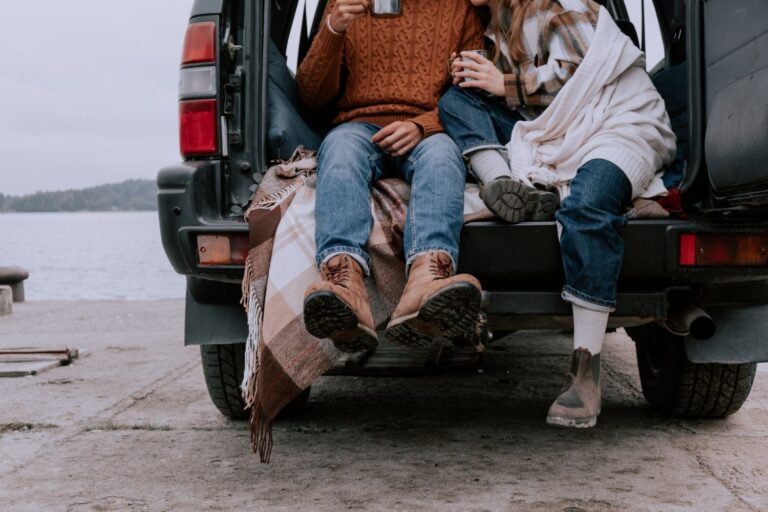 A couple enjoys a warm drink while sitting in an SUV trunk by a serene lakeside